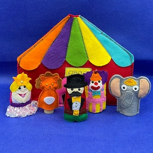 Può includere: Un set di burattini per dita in feltro a tema circense. I burattini includono un clown, un leone, un direttore di circo, una principessa e un elefante. I burattini sono davanti a una tenda da circo in feltro.