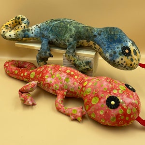 Può includere: Due lucertole di peluche fatte a mano, una blu e verde con macchie gialle e l'altra rossa con macchie gialle. Entrambe le lucertole hanno occhi neri con centri bianchi.