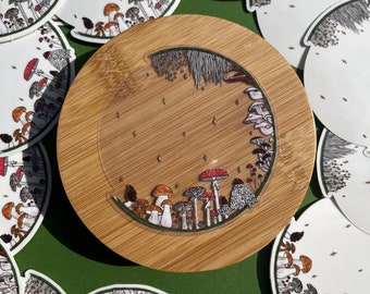 May include: A collection of round stickers featuring a whimsical illustration of mushrooms in a forest setting. The stickers are arranged on a wooden surface with a green background.