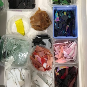 Op de afbeelding: Een verzameling kleine glazen stukjes in verschillende kleuren en vormen, georganiseerd in doorzichtige plastic containers. De stukjes zijn in rijen gerangschikt, waarbij elke container een andere kleur of type glas bevat.