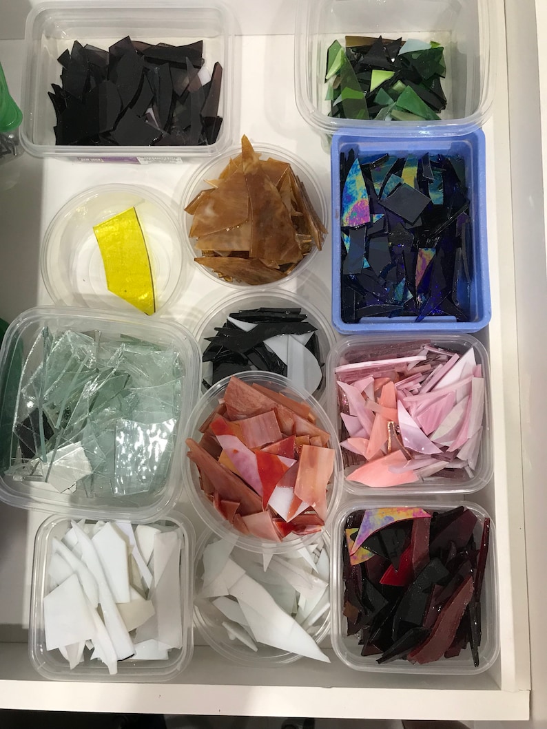 Op de afbeelding: Een verzameling kleine glazen stukjes in verschillende kleuren en vormen, georganiseerd in doorzichtige plastic containers. De stukjes zijn in rijen gerangschikt, waarbij elke container een andere kleur of type glas bevat.