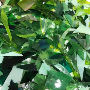 May include: A large pile of green glass shards, some with a slight blue tint, are scattered on a white surface. The glass pieces are various shapes and sizes.