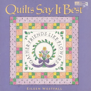 May include: A purple book cover with the title "Quilts Say It Best" in yellow lettering. The cover features a quilt block with a wreath of flowers and the text "Gather Friends Like Flowers".