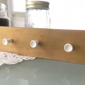 May include: A wooden wall-mounted coat rack with three white ceramic hooks. The rack is painted a light gold color.