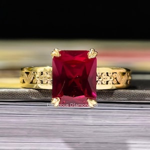 Könnte beinhalten: Ein goldener Ring mit einem großen, rechteckigen, roten Edelstein. Das Ringband weist ein kompliziertes Blumenmuster auf. Der Ring liegt auf einem Buch mit dunklem Hintergrund. Der Ring ist ein Schmuckstück.