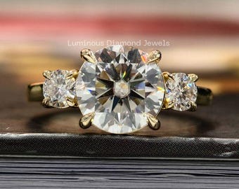 Può includere: Un anello di fidanzamento in oro con una grande pietra centrale rotonda e trasparente e due pietre laterali più piccole. L'anello è appoggiato su una superficie scura, con le parole "Luminous Diamond Jewels" visibili.