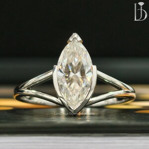 Könnte beinhalten: Ein Ring mit einem Diamanten im Marquiseschliff und einem silbernen Band. Der Diamant ist in einer Lünettenfassung gefasst, und das Band hat ein geteiltes Schaftdesign. Der Ring wird auf einer dunklen Oberfläche präsentiert. Der Text "Luminous Diamond Jewels" ist sichtbar.