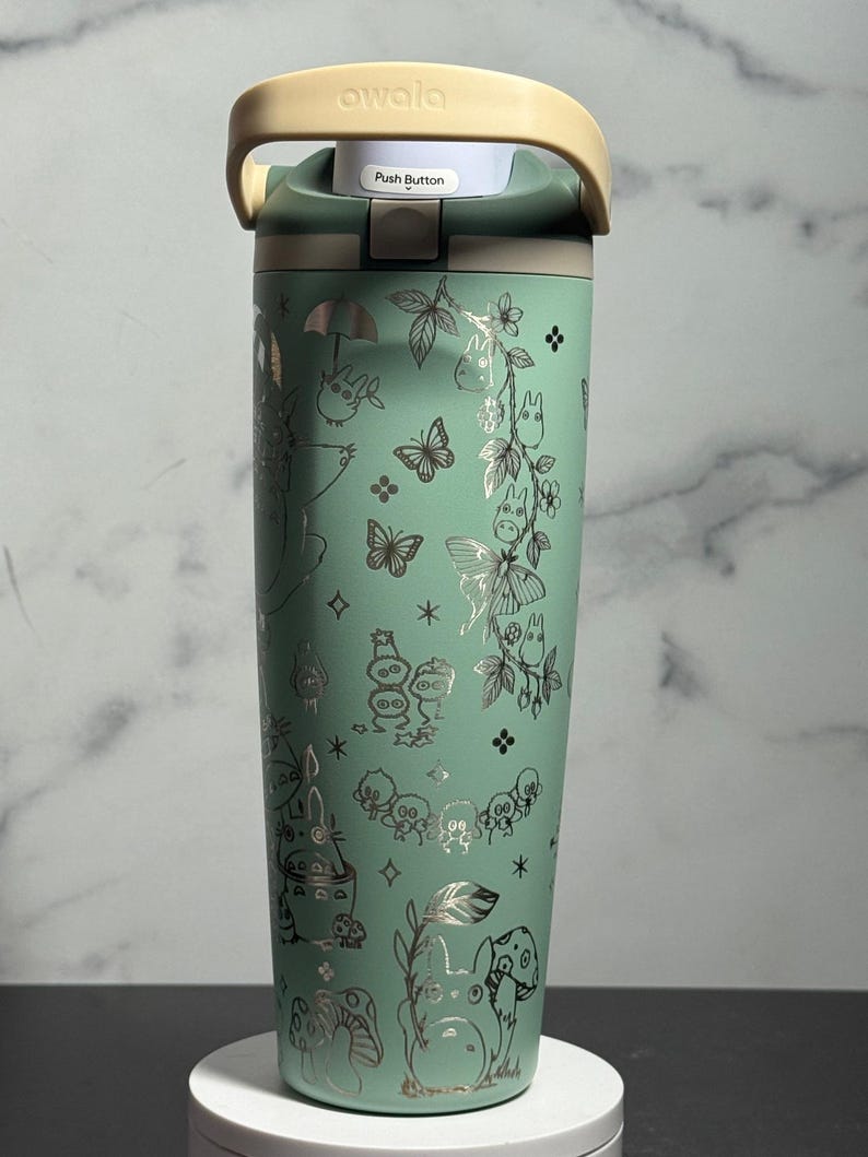 May include: A mint green Owala water bottle with a beige handle and lid. The bottle has silver illustrations of butterflies, mushrooms, and cartoon characters. The lid has the text "Push Button" and the brand name "owala".