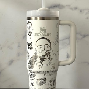 May include: White Stanley tumbler with a handle, straw, and lid. The tumbler features black illustrations and text, including the Stanley logo, portraits, and phrases like "Let's get lost in the clouds."