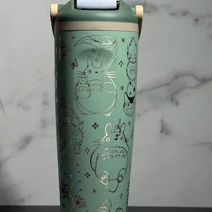 May include: A teal insulated tumbler with silver illustrations of characters from "My Neighbor Totoro." The tumbler has a flip-top lid and a handle. The design includes butterflies, stars, and floral accents. The tumbler is approximately 20 cm tall.