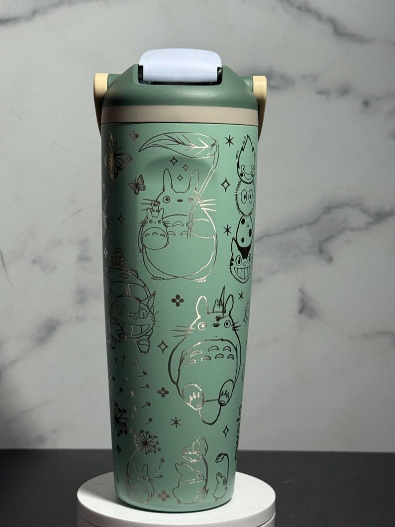 May include: A teal insulated tumbler with silver illustrations of characters from "My Neighbor Totoro." The tumbler has a flip-top lid and a handle. The design includes butterflies, stars, and floral accents. The tumbler is approximately 20 cm tall.