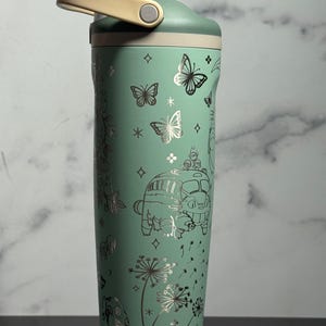 May include: A mint green insulated water bottle with a light beige handle and lid. The bottle is decorated with silver illustrations of butterflies, flowers, and a cartoon bus. The bottle is designed to keep drinks cold or hot.