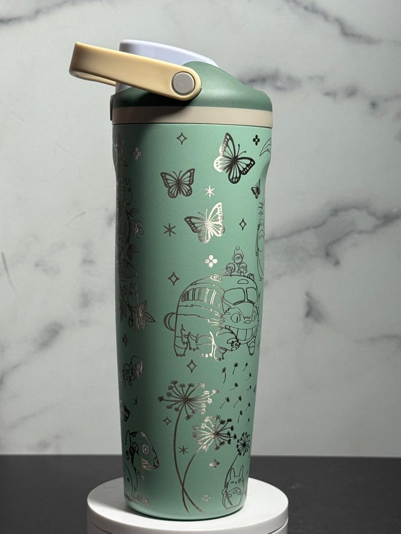 May include: A mint green insulated water bottle with a light beige handle and lid. The bottle is decorated with silver illustrations of butterflies, flowers, and a cartoon bus. The bottle is designed to keep drinks cold or hot.