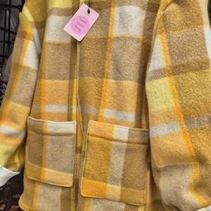 May include: A yellow, tan, and white checked jacket with two large front pockets. The jacket has a collarless design and a tag that reads "Emma Makes Things". The jacket is made of a thick, textured fabric.