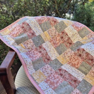 May include: A small, handmade quilt with a patchwork design. The quilt features squares of fabric in various floral patterns and solid colours, including pink, grey, and yellow. The quilt is trimmed with a pink border and is draped over a chair.