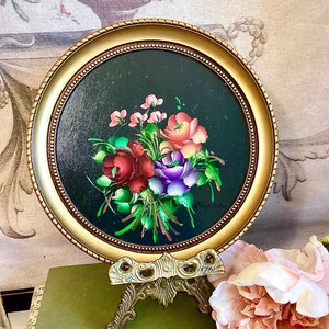 May include: A round, gold-framed painting of a colourful floral bouquet on a black background. The painting is on a small, gold-coloured easel.