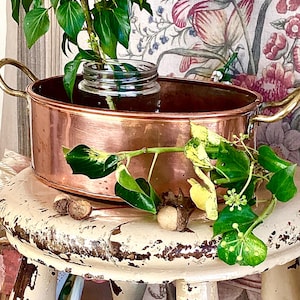 May include: A copper oval planter with brass handles contains a small glass jar with a green plant. The planter rests on a distressed white stool, with green ivy and acorns nearby. A floral patterned backdrop is visible.
