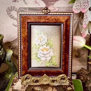 May include: A framed floral painting featuring two white peony flowers with yellow centres and green leaves. The painting is set in a brown wooden frame with a gold beaded border and a decorative brass stand.