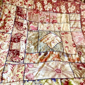 May include: A patchwork quilt featuring floral and paisley designs in red, cream, and pink hues. The quilt is constructed from rectangular and square fabric pieces, joined with dark blue stitching. The overall aesthetic blends vintage and rustic elements.