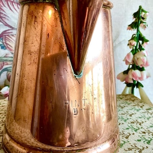 May include: A copper pint jug with a spout. The jug has a wide base and tapers upwards. The word "PINT" is stamped on the front. The jug is made of copper and has a shiny, reflective surface.