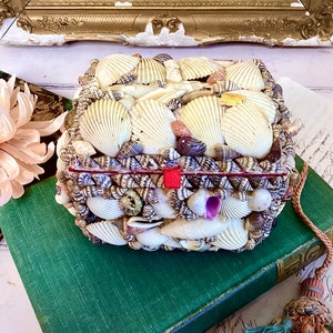 May include: A small, rectangular box entirely covered in a mosaic of various seashells. The box has a red ribbon tied around it and is sitting on top of a green book.