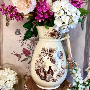 May include: A vintage cream-coloured ceramic pitcher with a brown floral design and a tassel handle. The pitcher is filled with a bouquet of pink, white and purple flowers.