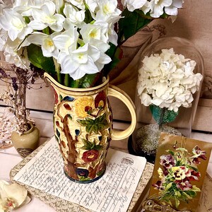 May include: A vintage ceramic jug with a floral design in shades of yellow, red, and green. The jug is filled with white flowers and sits on a stack of books with a postcard on a decorative stand.