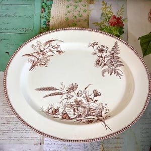May include: Oval, cream-coloured serving platter with a brown floral and fauna design. The rim has a detailed brown border. The platter features illustrations of flowers, wheat, rabbits, and a landscape scene. A vintage-style piece.
