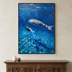Op de afbeelding: Een rechthoekige wandkunstprint met een olieverfschilderij van een zeemeermin met een blauwe staart die onder water naast een lamantijn zwemt. De scène is weergegeven in diepe blauwtinten met licht dat reflecteert op de zanderige zeebodem.