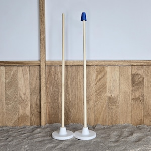 Puede incluir: Dos mástiles de bandera de madera con bases blancas. Uno es liso, el otro tiene una parte superior azul. Los postes se alzan sobre una superficie texturizada, contra un fondo de paneles de madera y una pared blanca.