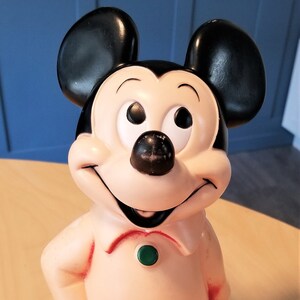 May include: A vintage Mickey Mouse figurine, featuring black ears, a black nose, and a smiling face. The figure wears a light peach-colored shirt, blue pants, and black shoes, standing on a green base. A classic Disney collectible.