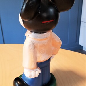 May include: A vintage Mickey Mouse figurine, viewed from the side. The figure has black ears and head, a peach-colored shirt, blue pants, and a green base. The coin slot is visible. The figurine is standing on a light-colored surface.