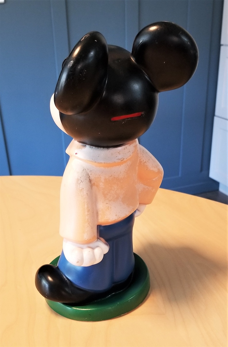 May include: A vintage Mickey Mouse figurine, viewed from the side. The figure has black ears and head, a peach-colored shirt, blue pants, and a green base. The coin slot is visible. The figurine is standing on a light-colored surface.
