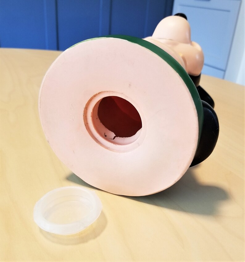 May include: A vintage Snoopy ceramic coin bank, viewed from the bottom. The bank features a light pink base, a green band, and black accents. A clear plastic stopper sits beside the bank. The coin slot is visible, and the interior is red.