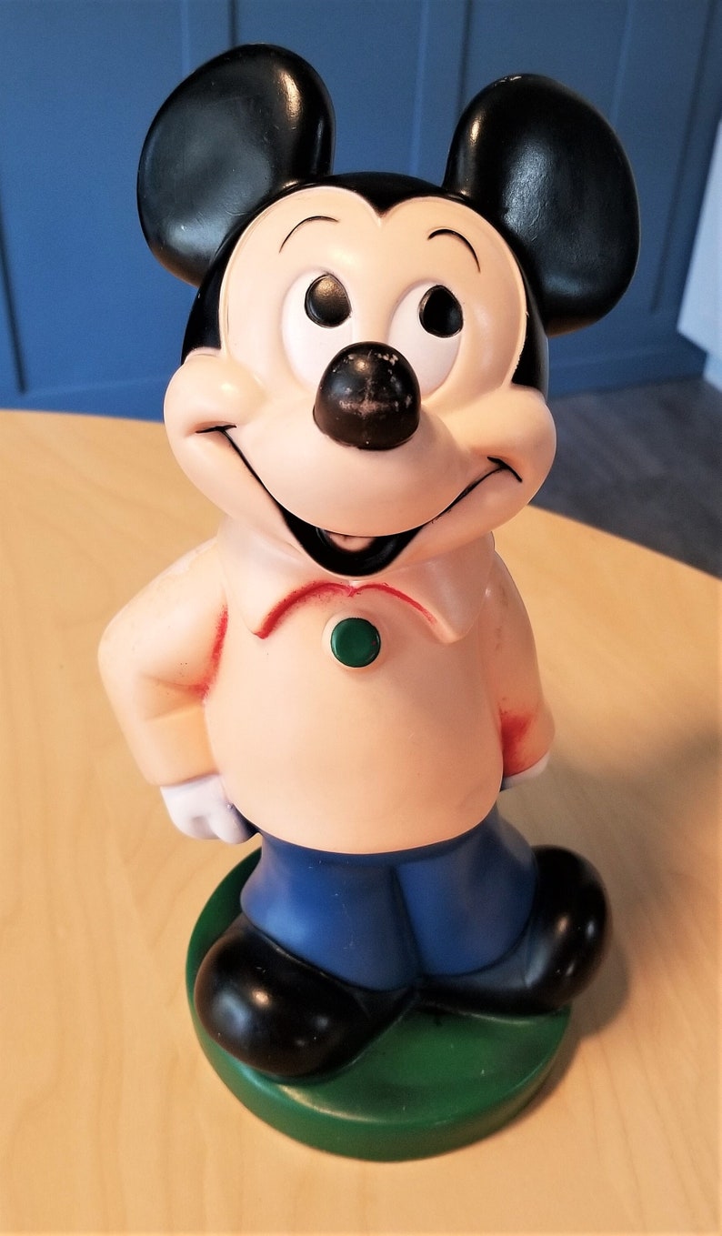 May include: A vintage Mickey Mouse figurine, featuring black ears, a black nose, and a smiling face. The figure wears a light peach-colored shirt, blue pants, and black shoes, standing on a green base. A classic Disney collectible.