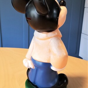 May include: A vintage Mickey Mouse figurine, featuring a black head and ears, a tan body, blue pants, and black shoes. The figurine is standing on a green base. The overall design is a classic cartoon character.