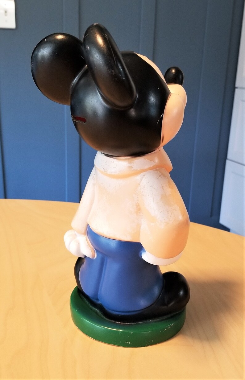 May include: A vintage Mickey Mouse figurine, featuring a black head and ears, a tan body, blue pants, and black shoes. The figurine is standing on a green base. The overall design is a classic cartoon character.