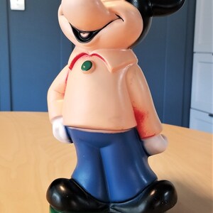 May include: A vintage Mickey Mouse figurine. The figure has a peach-colored shirt, blue pants, and black shoes. The base is green. The figure has black ears and a smiling face.