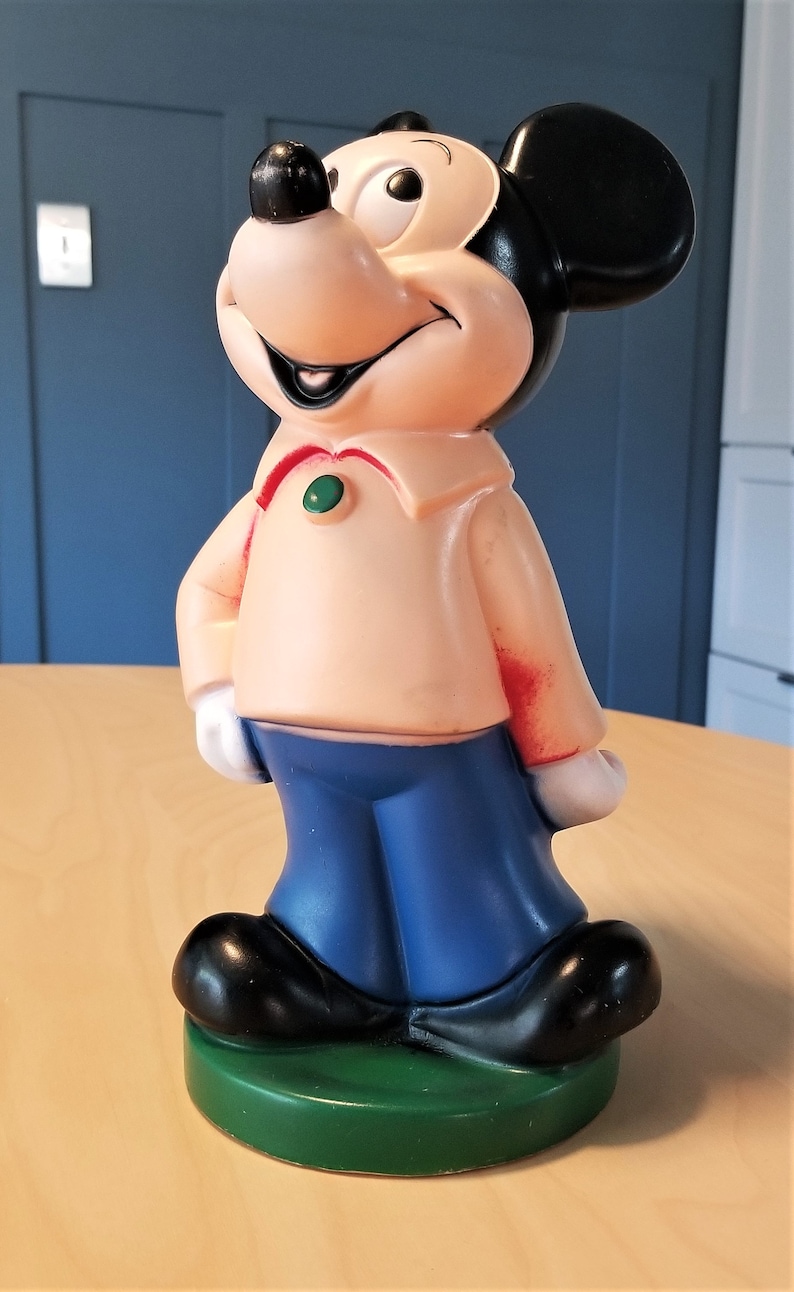 May include: A vintage Mickey Mouse figurine. The figure has a peach-colored shirt, blue pants, and black shoes. The base is green. The figure has black ears and a smiling face.