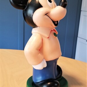 May include: A vintage Mickey Mouse figurine. The figure has a black head and ears, a light pink body, blue pants, and black shoes. It stands on a green base. The figurine is a classic Disney collectible.