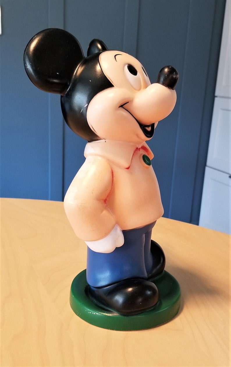 May include: A vintage Mickey Mouse figurine. The figure has a black head and ears, a light pink body, blue pants, and black shoes. It stands on a green base. The figurine is a classic Disney collectible.