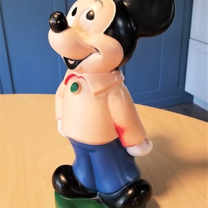 May include: A vintage Mickey Mouse figurine. The figure is primarily beige, black, and blue, with a green base. Mickey is wearing a beige shirt, blue pants, and black shoes. His iconic black ears are prominent.