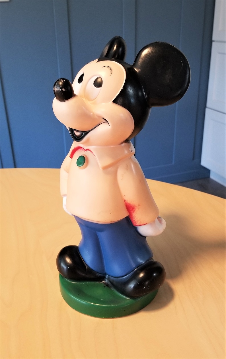 May include: A vintage Mickey Mouse figurine. The figure is primarily beige, black, and blue, with a green base. Mickey is wearing a beige shirt, blue pants, and black shoes. His iconic black ears are prominent.