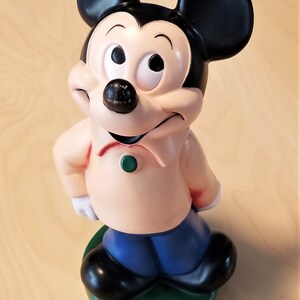 May include: A Mickey Mouse figurine, featuring black ears, a black nose, and a peach-colored shirt with a green button. The figure has blue pants and black shoes, standing on a green base. A classic Disney collectible.
