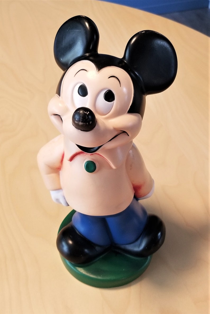 May include: A Mickey Mouse figurine, featuring black ears, a black nose, and a peach-colored shirt with a green button. The figure has blue pants and black shoes, standing on a green base. A classic Disney collectible.