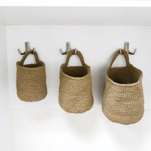 May include: Three brown crocheted hanging baskets with rope handles are hanging on hooks on a white wall.