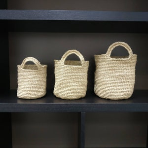 May include: Three beige crocheted baskets of varying sizes, with handles, are displayed on a black shelf.