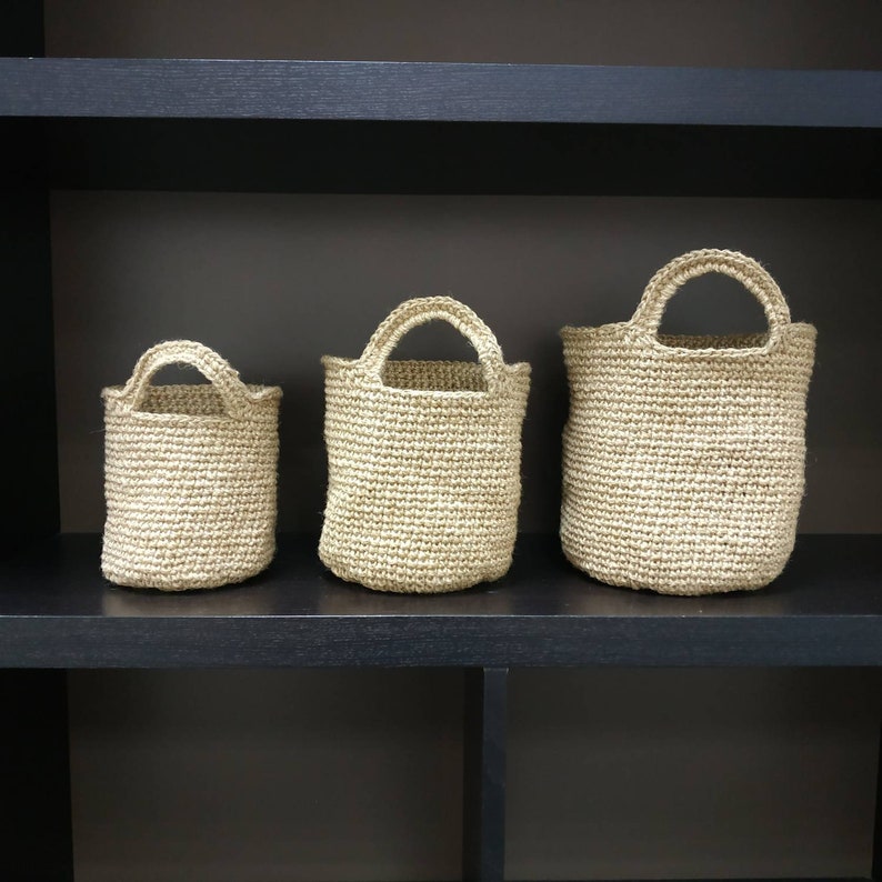 May include: Three beige crocheted baskets of varying sizes, with handles, are displayed on a black shelf.