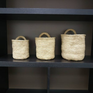 May include: Three woven baskets of varying sizes, made of natural fibers, are displayed on a black shelf. The baskets have a natural, light brown color and are made with a tight weave.