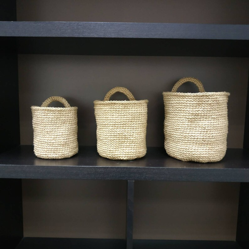 May include: Three woven baskets of varying sizes, made of natural fibers, are displayed on a black shelf. The baskets have a natural, light brown color and are made with a tight weave.